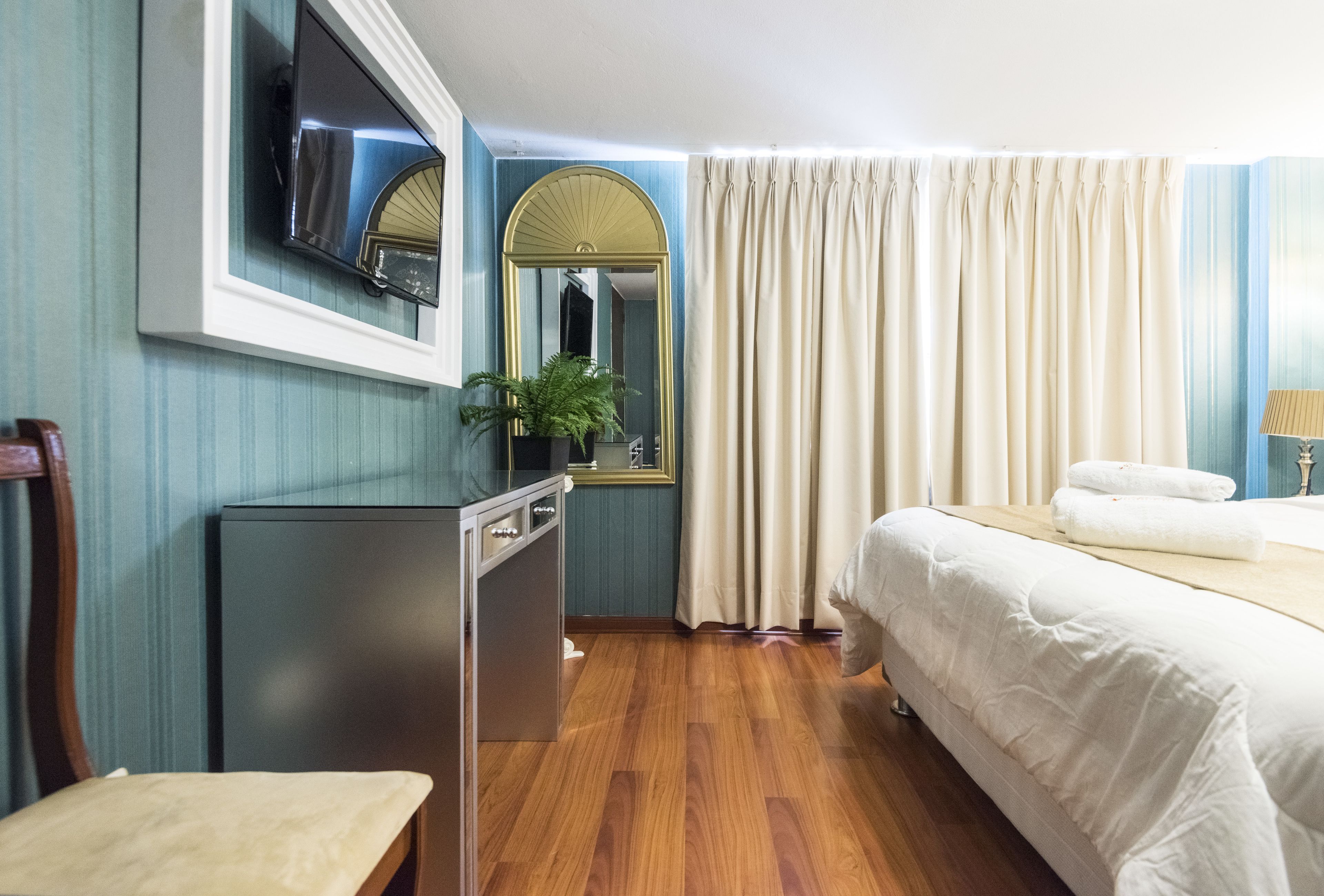 A hotel room with a bed, a desk, a chair, a TV, a mirror, a plant, and curtains.