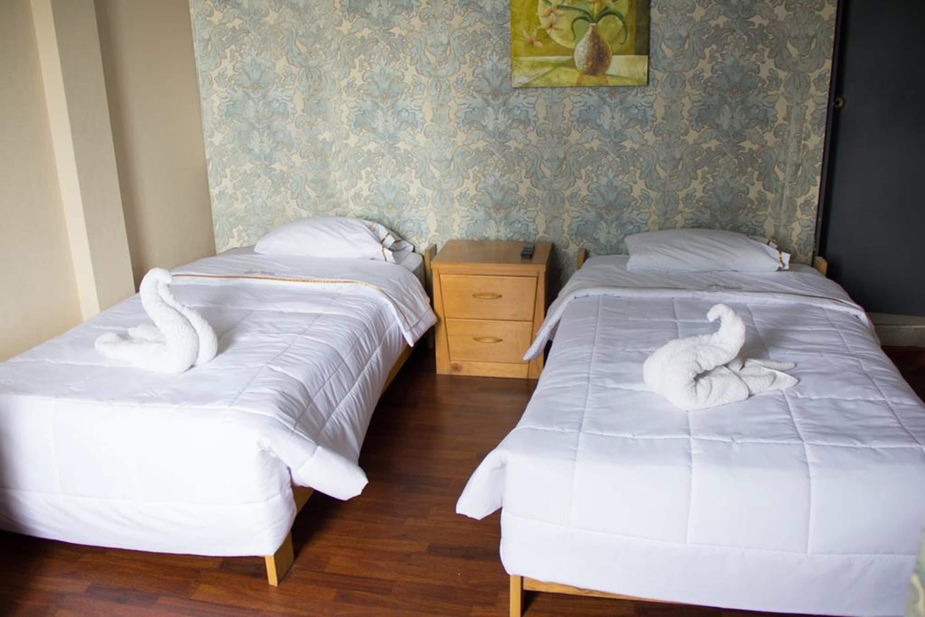 Habitación de hotel con dos camas individuales, cada una con un edredón blanco y una toalla con forma de cisne. Hay una mesita de noche de madera entre las camas.