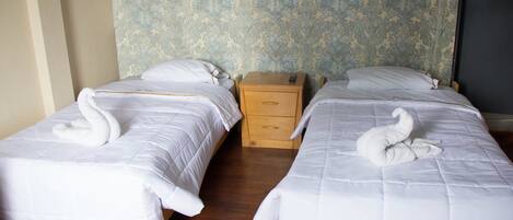 Habitación de hotel con dos camas individuales, cada una con un edredón blanco y una toalla con forma de cisne. Hay una mesita de noche de madera entre las camas.