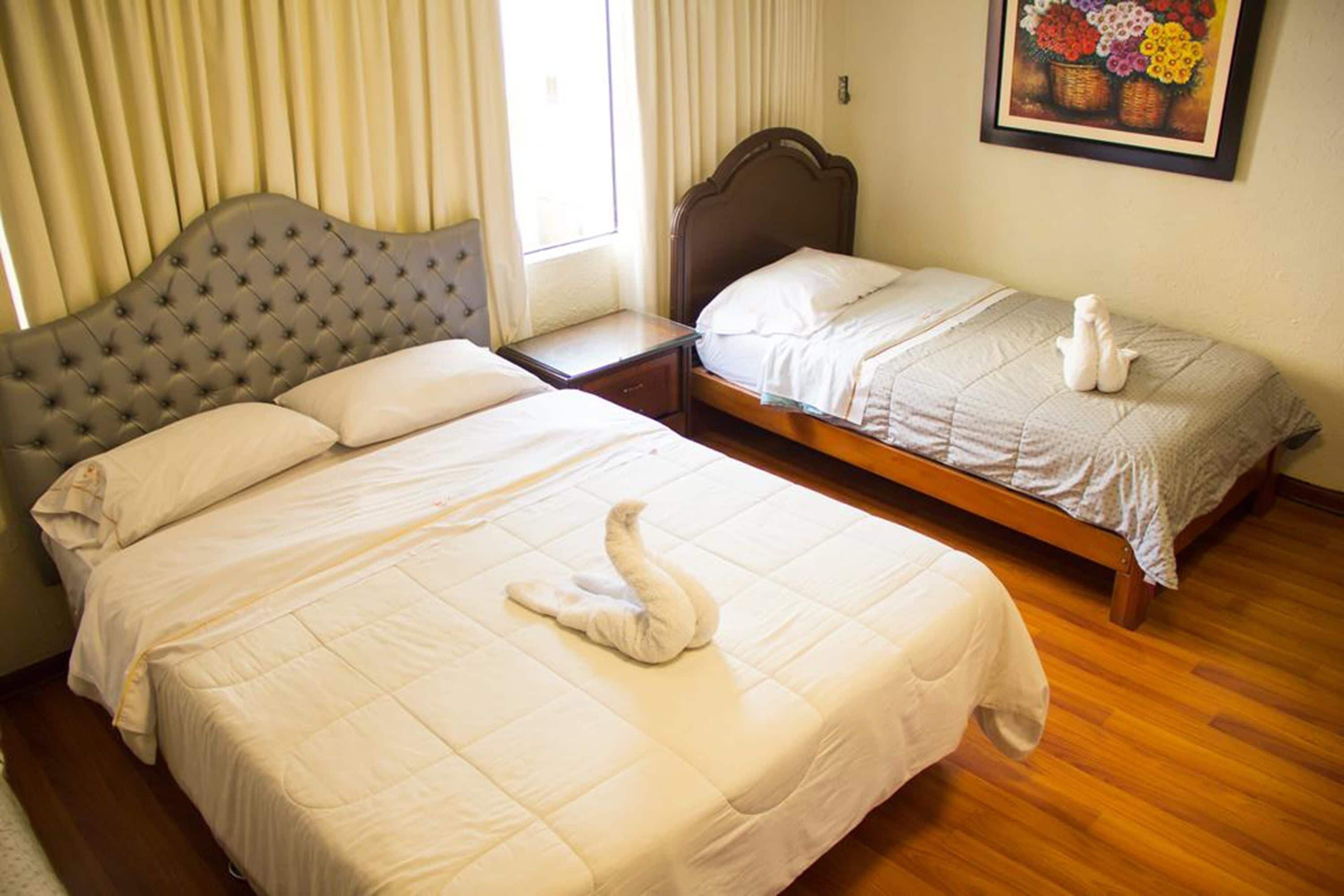 A hotel room with two beds, one larger and one smaller, both with white bedding. A swan-shaped towel is placed on the larger bed.