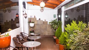 An outdoor patio with wicker chairs and round tables, surrounded by potted plants and a stone wall.