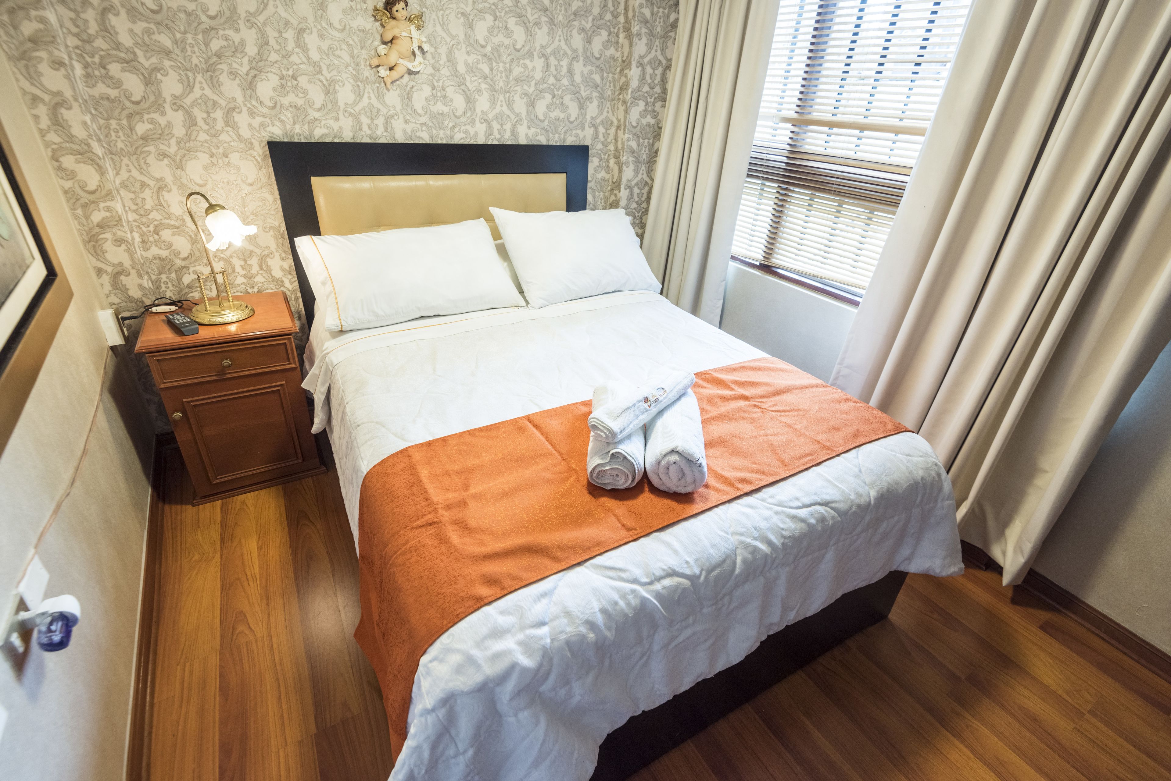 A neatly made bed with white linens, an orange blanket, and rolled towels on the bed. There is a nightstand with a lamp and a remote control beside the bed. The room has wooden flooring and a window with curtains.