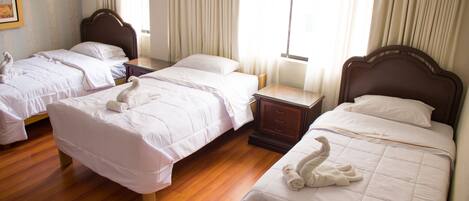 A hotel room with three single beds, wooden flooring, and a large window with curtains.