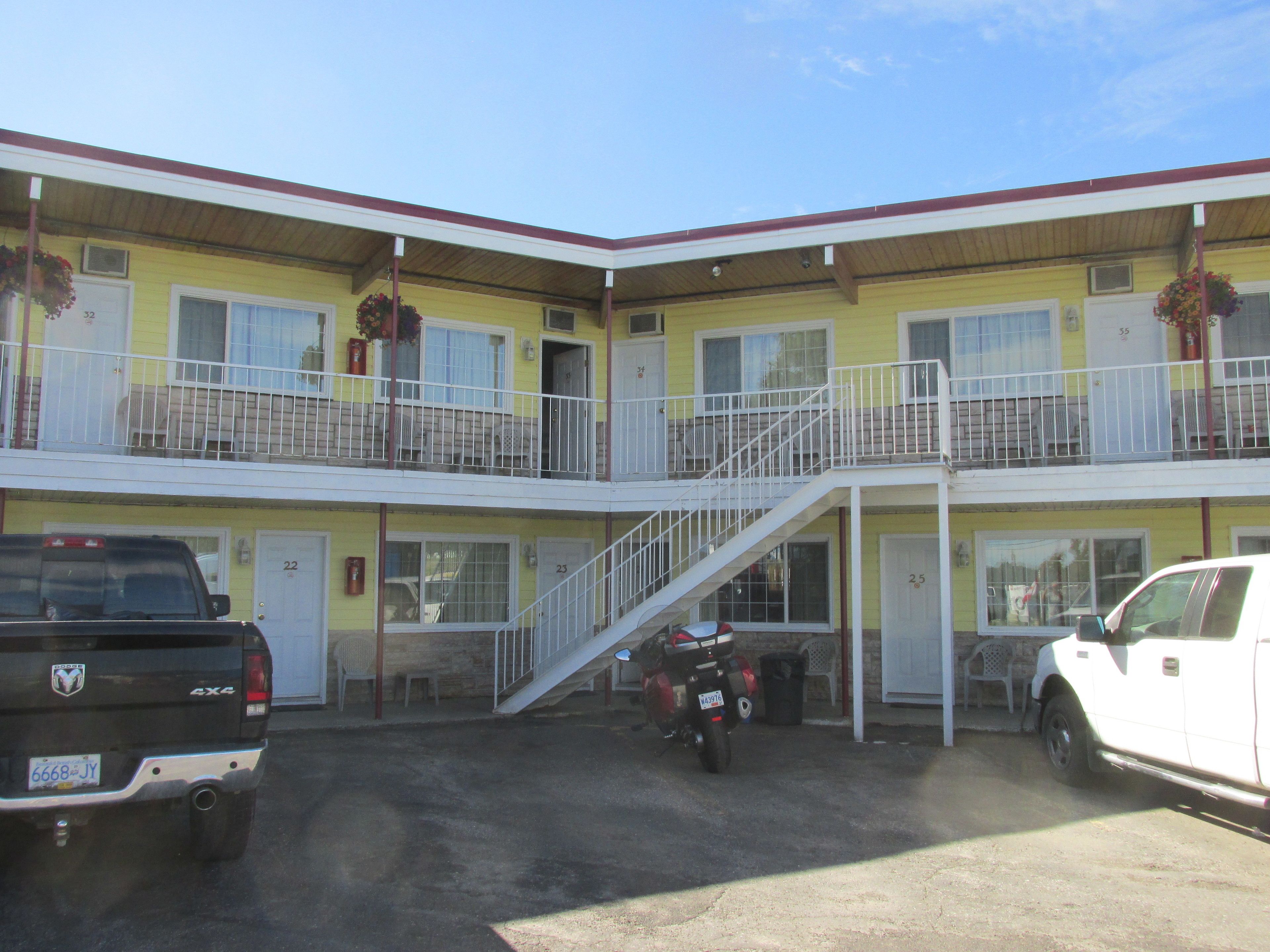 Um motel de dois andares com uma escadaria central, vários quartos e um estacionamento com dois veículos.