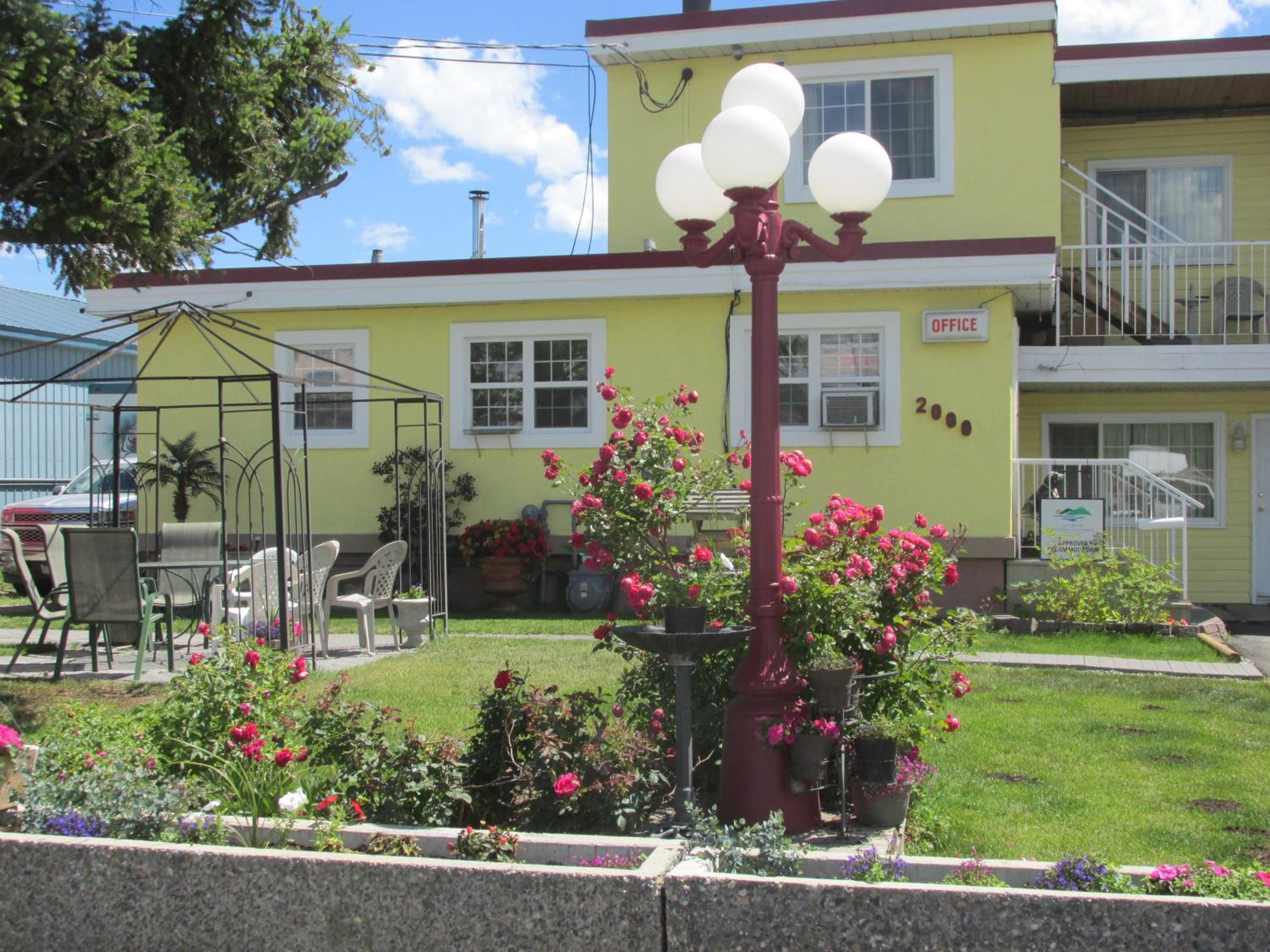 A yellow building with a sign indicating an office, a garden with flowers, and a metal structure with spherical lights.