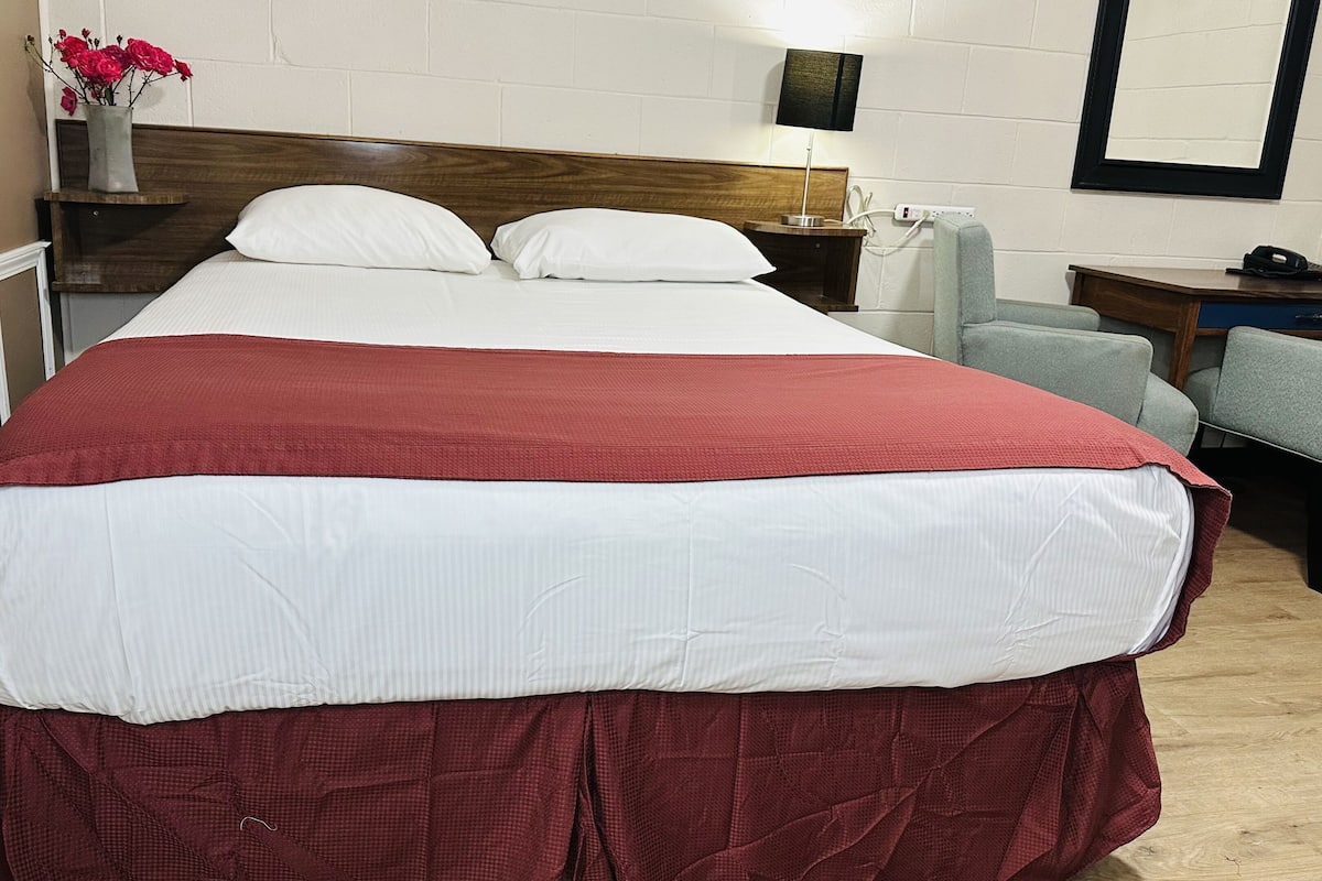 Quarto de hotel com uma cama grande, mesa de cabeceira com abajur, cadeira e escrivaninha com telefone.