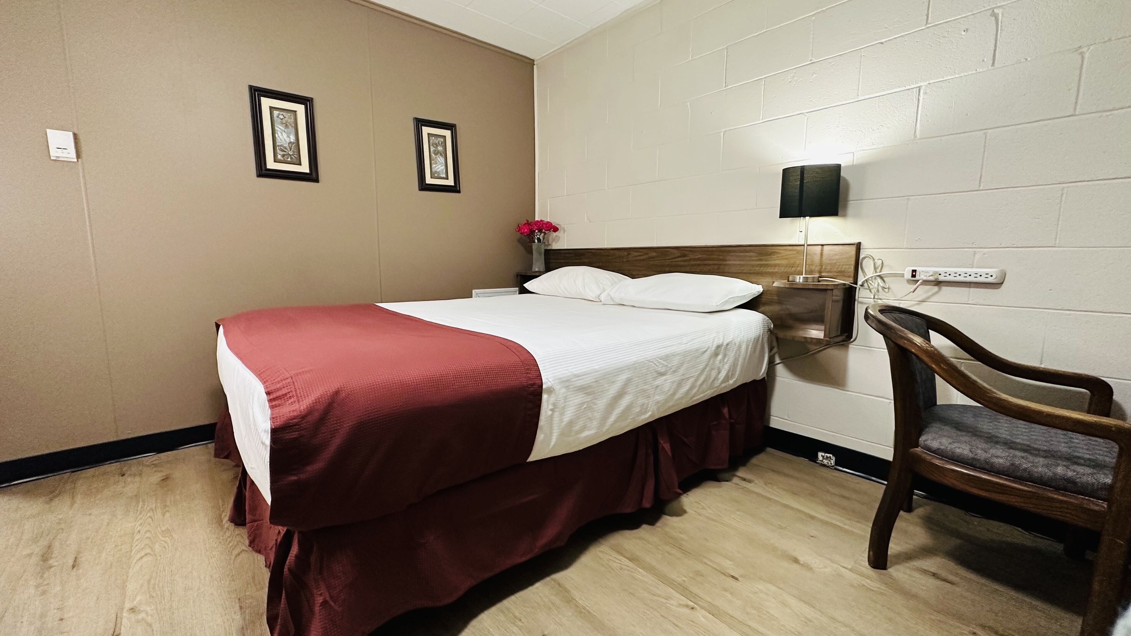 A hotel room with a bed, a nightstand with a lamp, a chair, and two framed pictures on the wall.