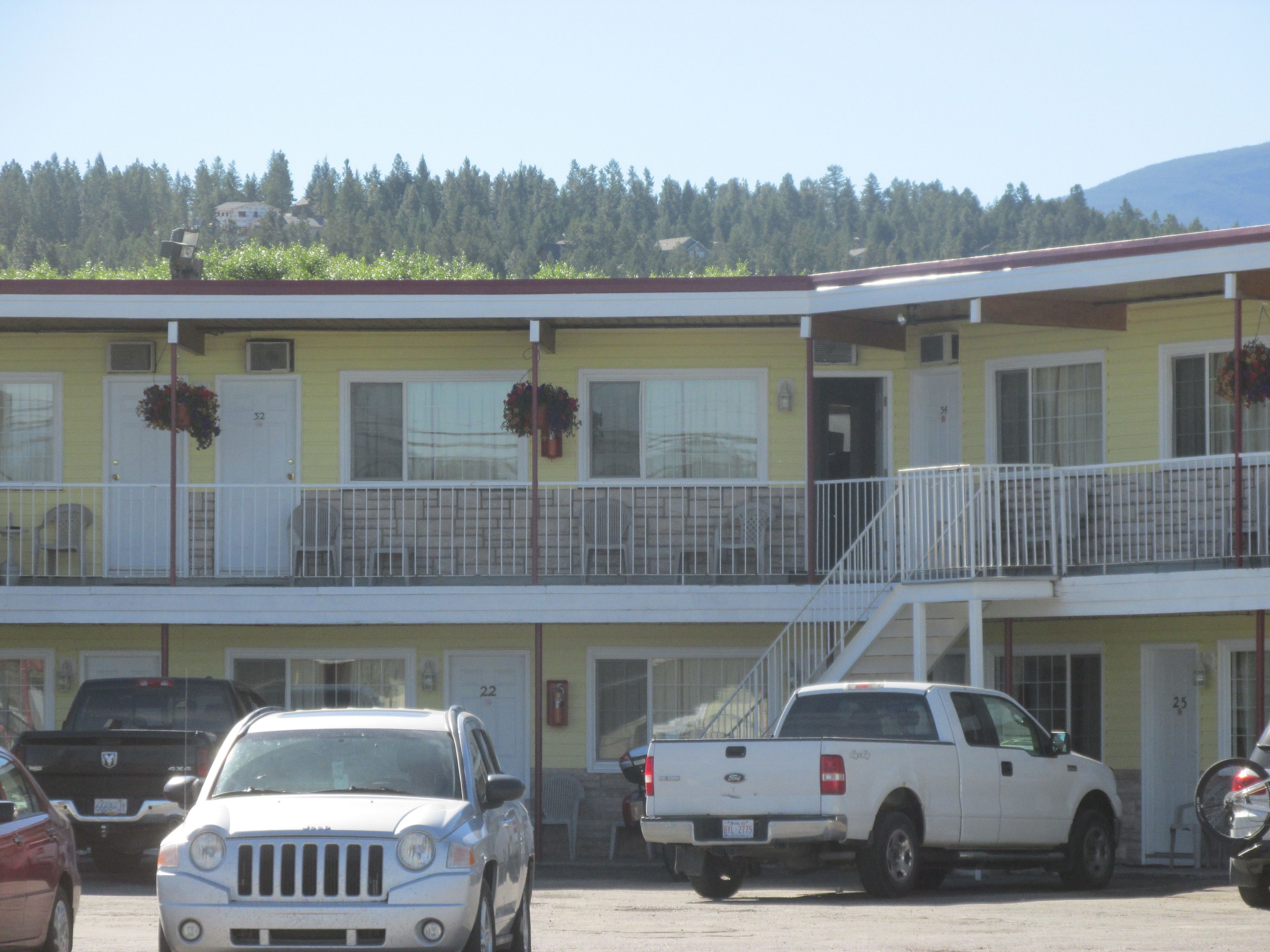 A two-story motel with a yellow facade, a balcony with white railings, and a parking lot with several vehicles.