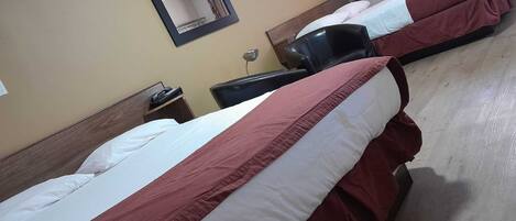 A hotel room with two beds, a nightstand, a chair, a mirror, and a wall-mounted lamp.