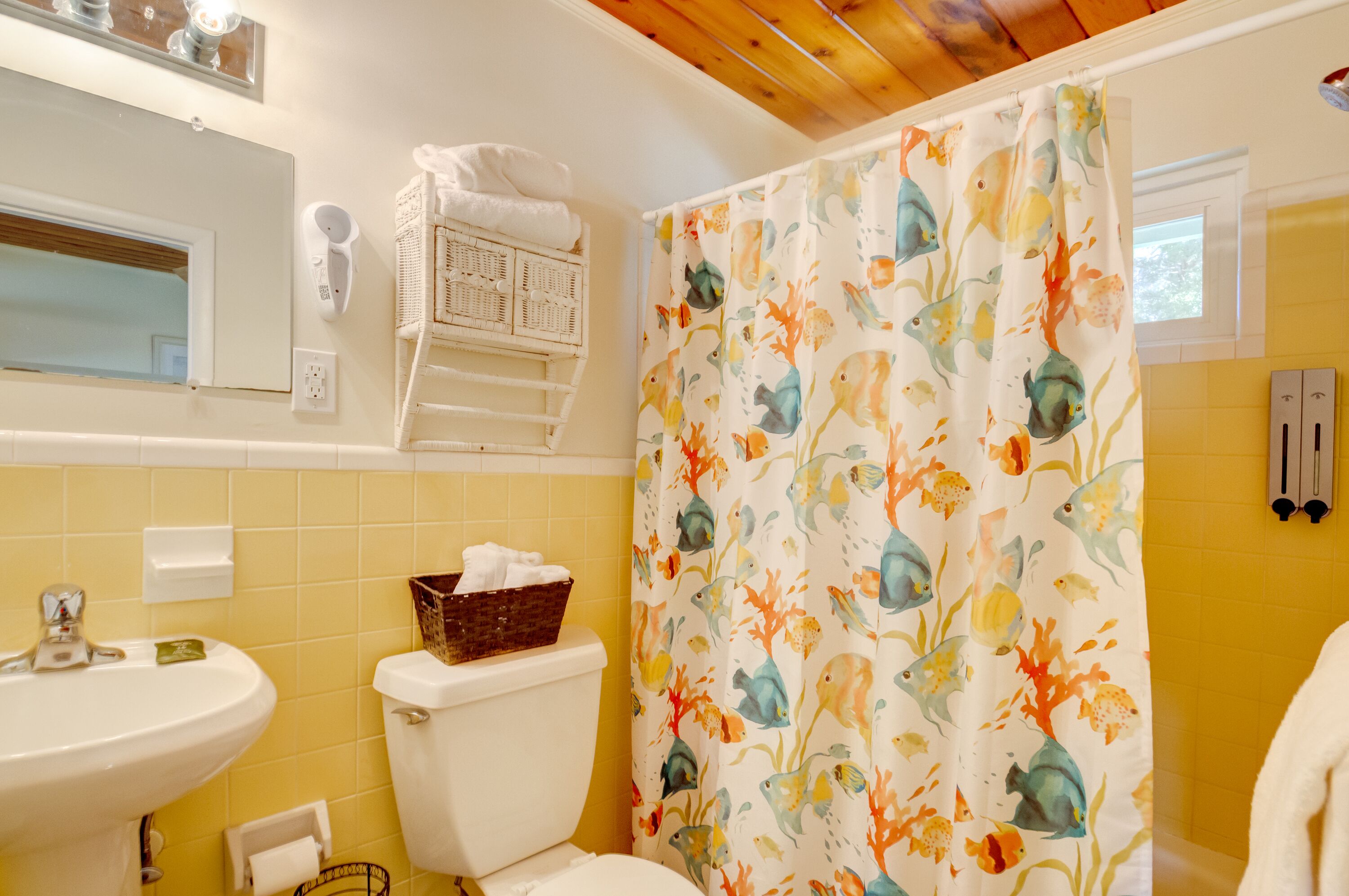 A bathroom with a yellow tiled wall, a white sink, a toilet, a shower curtain with a fish pattern, and a mirror above the sink.