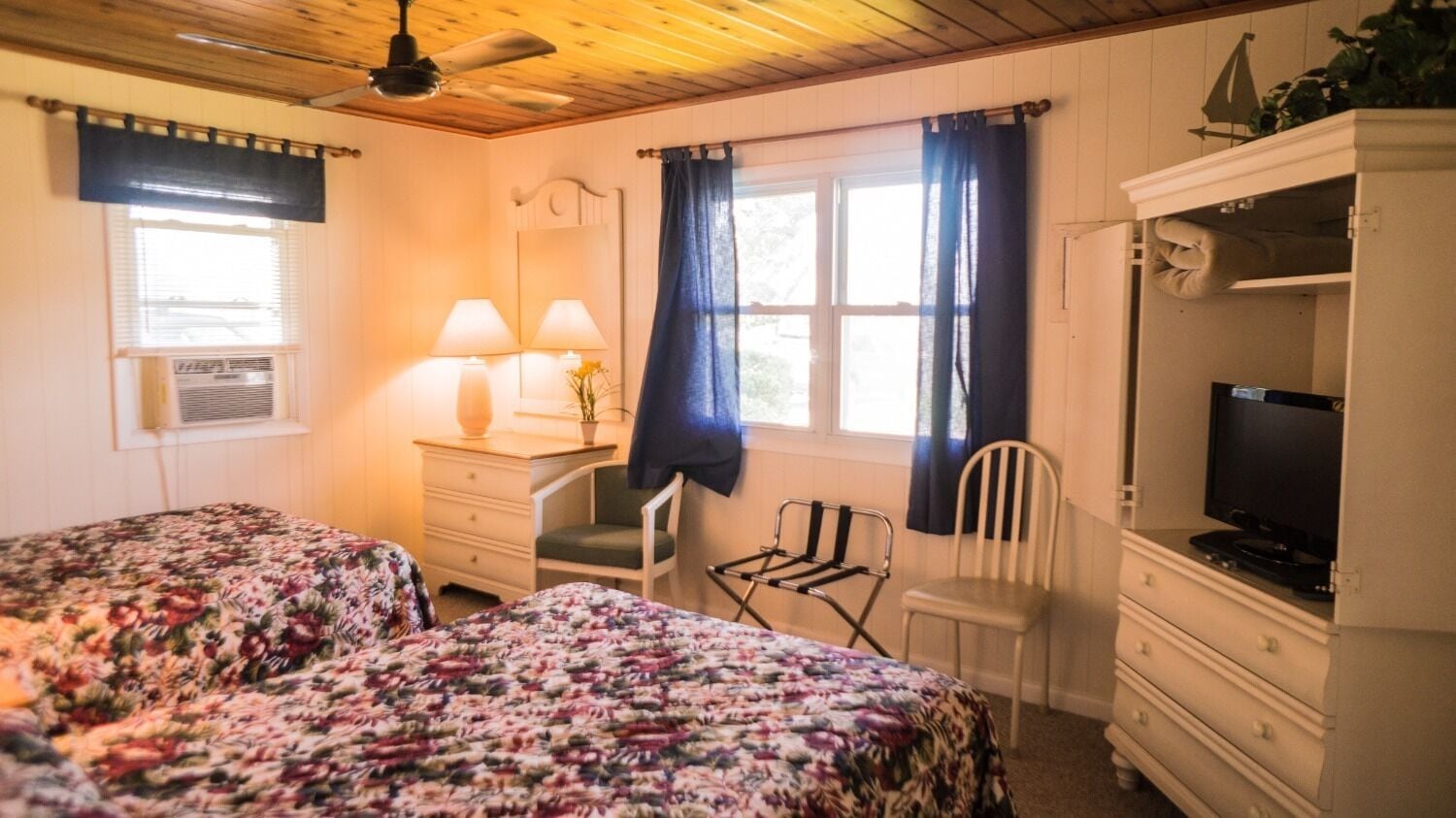A bedroom with two beds, a desk with a chair, a television, and a window with curtains.