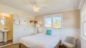 A bedroom with a bed, a chair, a bedside table with a lamp, a ceiling fan, a window with a view, and a door with towels hanging on it.