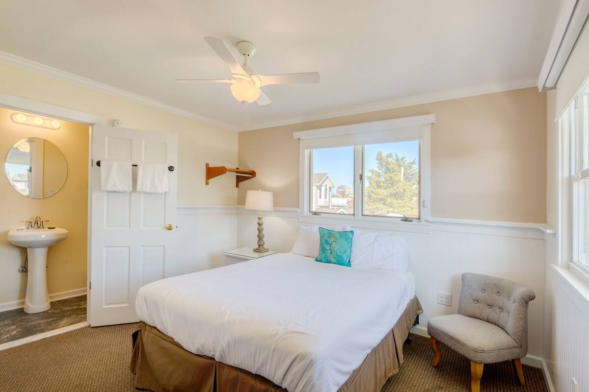 Quarto com cama, poltrona, mesa de cabeceira com abajur, ventilador de teto, janela com vista e porta com toalhas penduradas.