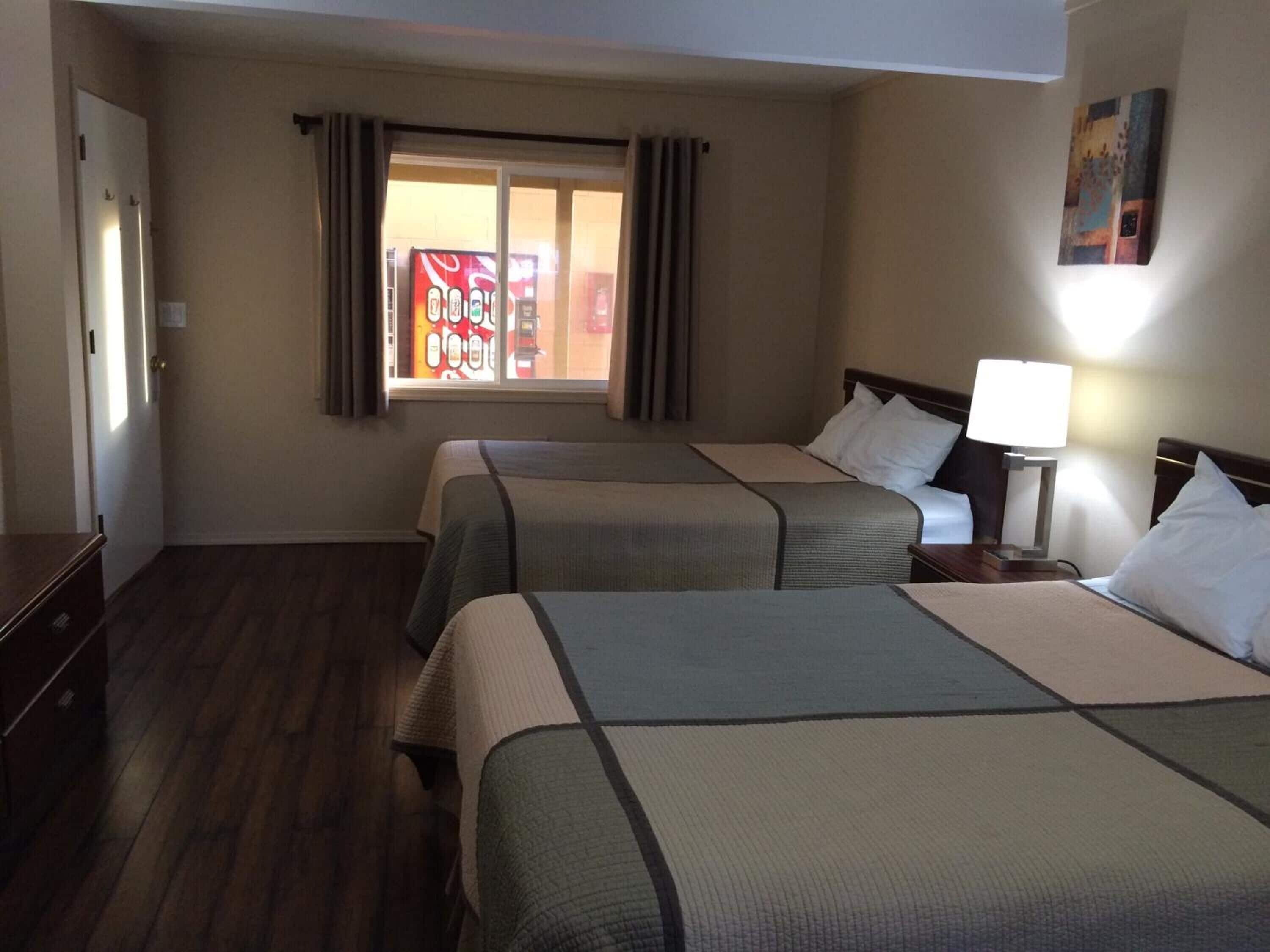 Habitación de hotel con dos camas, una mesita de noche con lámpara, una ventana con cortinas y una puerta.