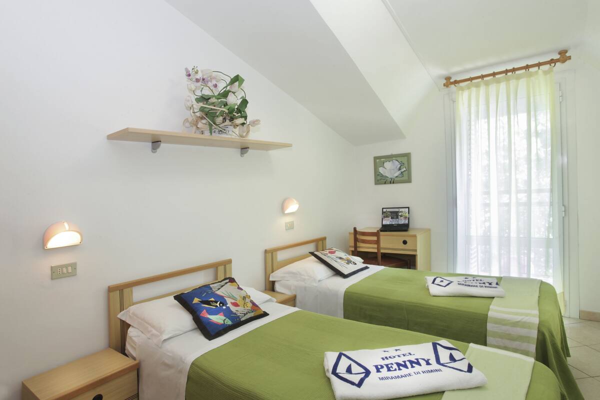 A hotel room with two beds, a desk, a chair, a shelf with plants, and a window with curtains.