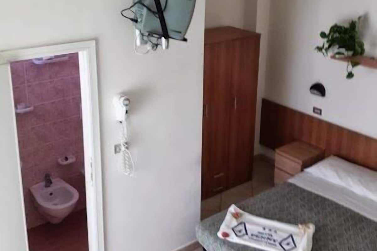 A small, compact room with a bed, a wooden wardrobe, a small table, and a wall-mounted shelf.