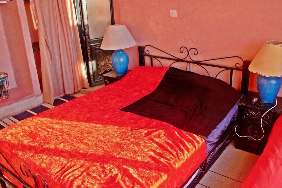 Ein Bett mit roter und schwarzer Bettdecke, zwei Nachttischlampen und ein kleiner Tisch mit einer Fernbedienung.