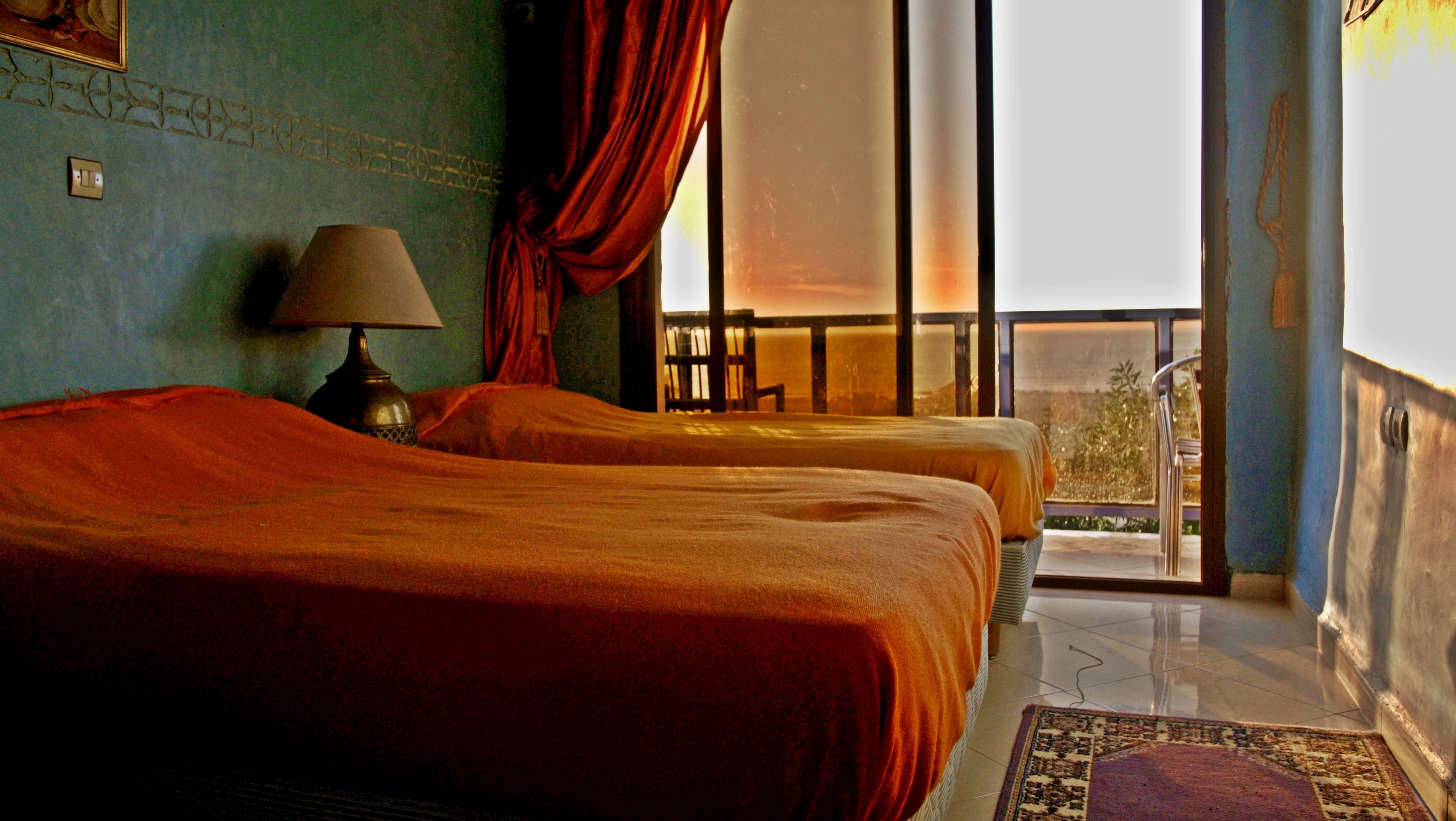 A hotel room with two beds, a lamp, a window with curtains, and a view of the outdoors.