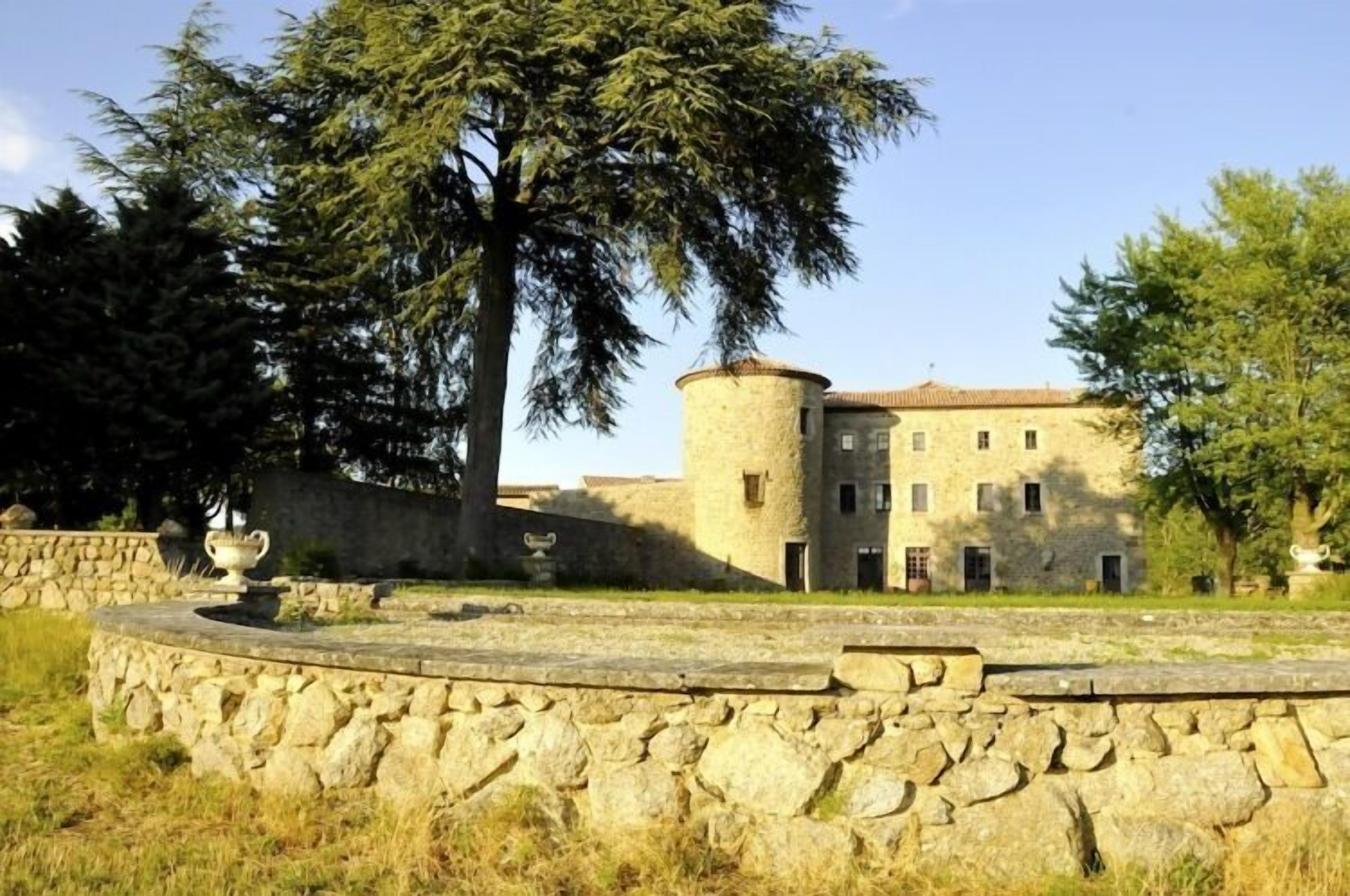 Un edificio de piedra con una torre redonda, rodeado por un muro de piedra y una plataforma circular de piedra con dos jarrones.