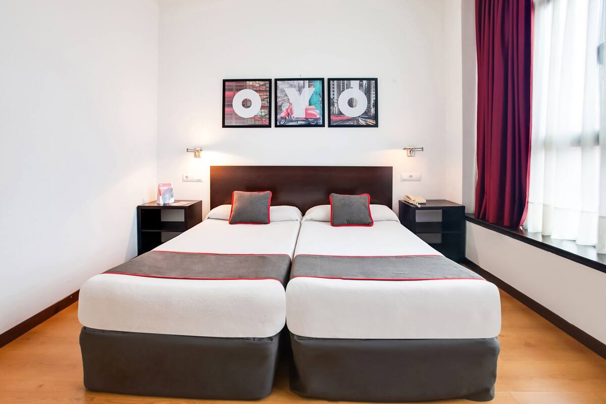 A double bed with a headboard, two bedside tables, and a window with red curtains.