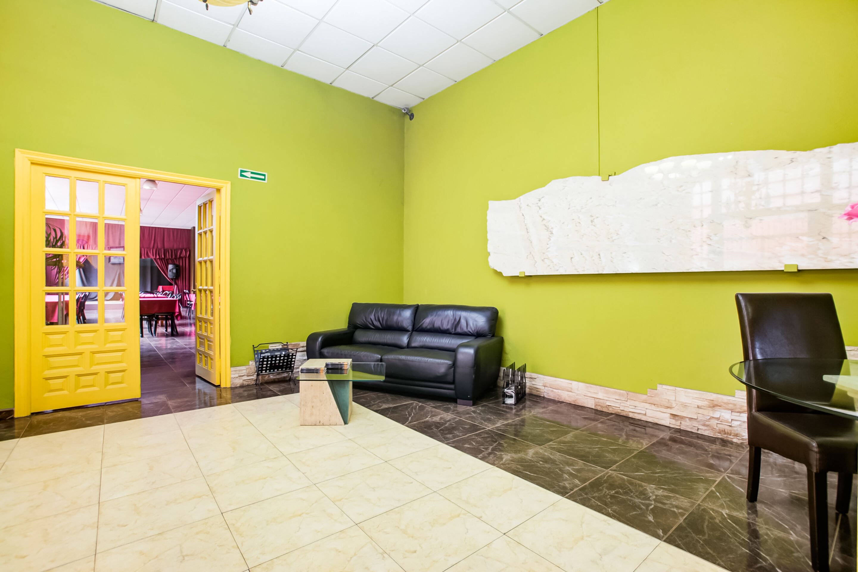 Un vestíbulo con una pared verde, un sofá de cuero negro, una mesa de vidrio y una puerta amarilla que da a un comedor.