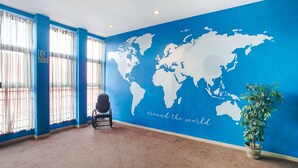 A room with a blue wall featuring a world map and the phrase "around the world." There is a wheelchair, a potted plant, and a window with curtains. - Parque Empresarial by Vivere Stays (Aviles)