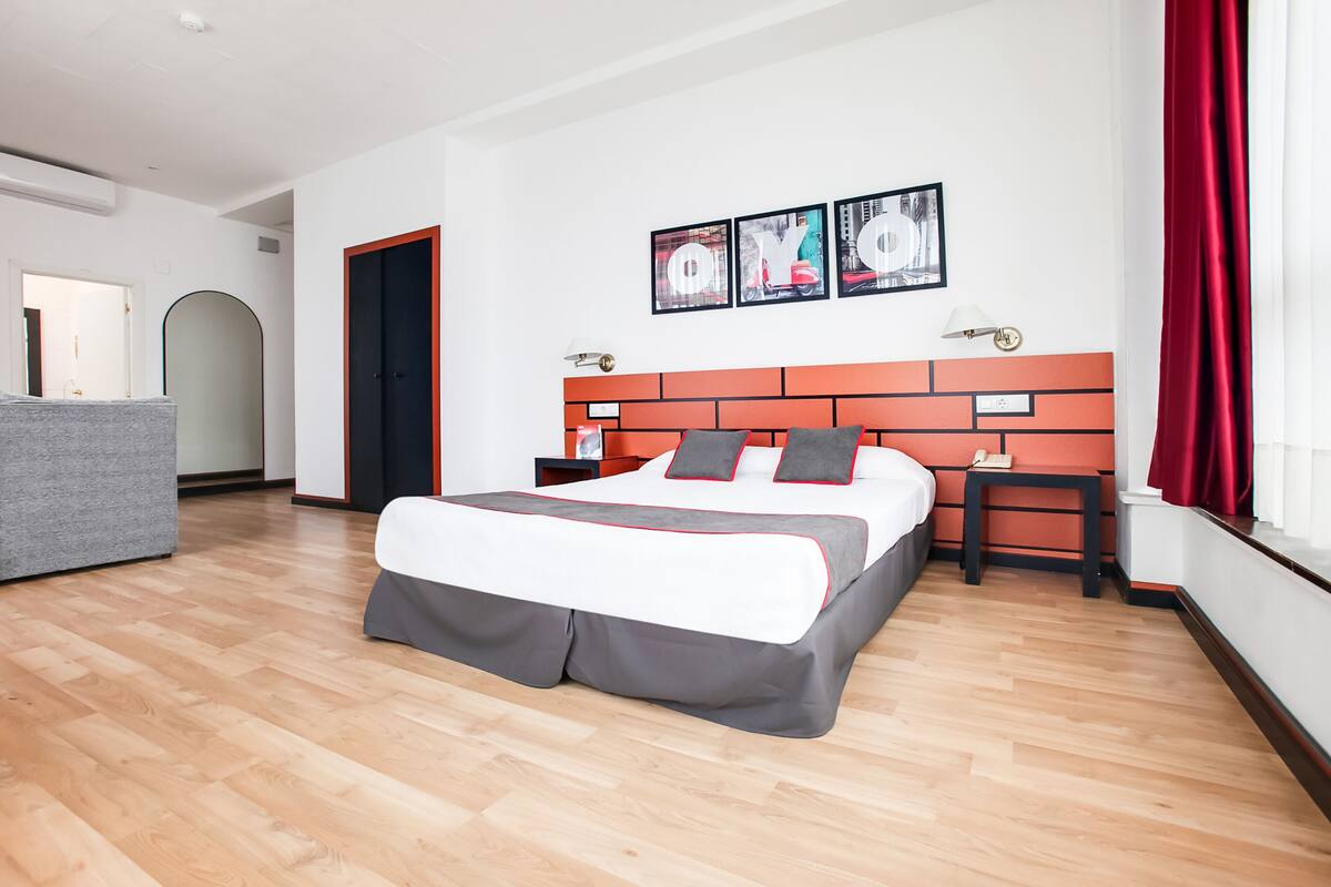 A modern hotel room with a large bed, a grey sofa, a red curtain, and a wall with three framed pictures.