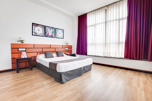 Una habitación de hotel moderna con una cama grande, una mesita de noche, una ventana con cortinas rojas y una obra de arte en la pared.