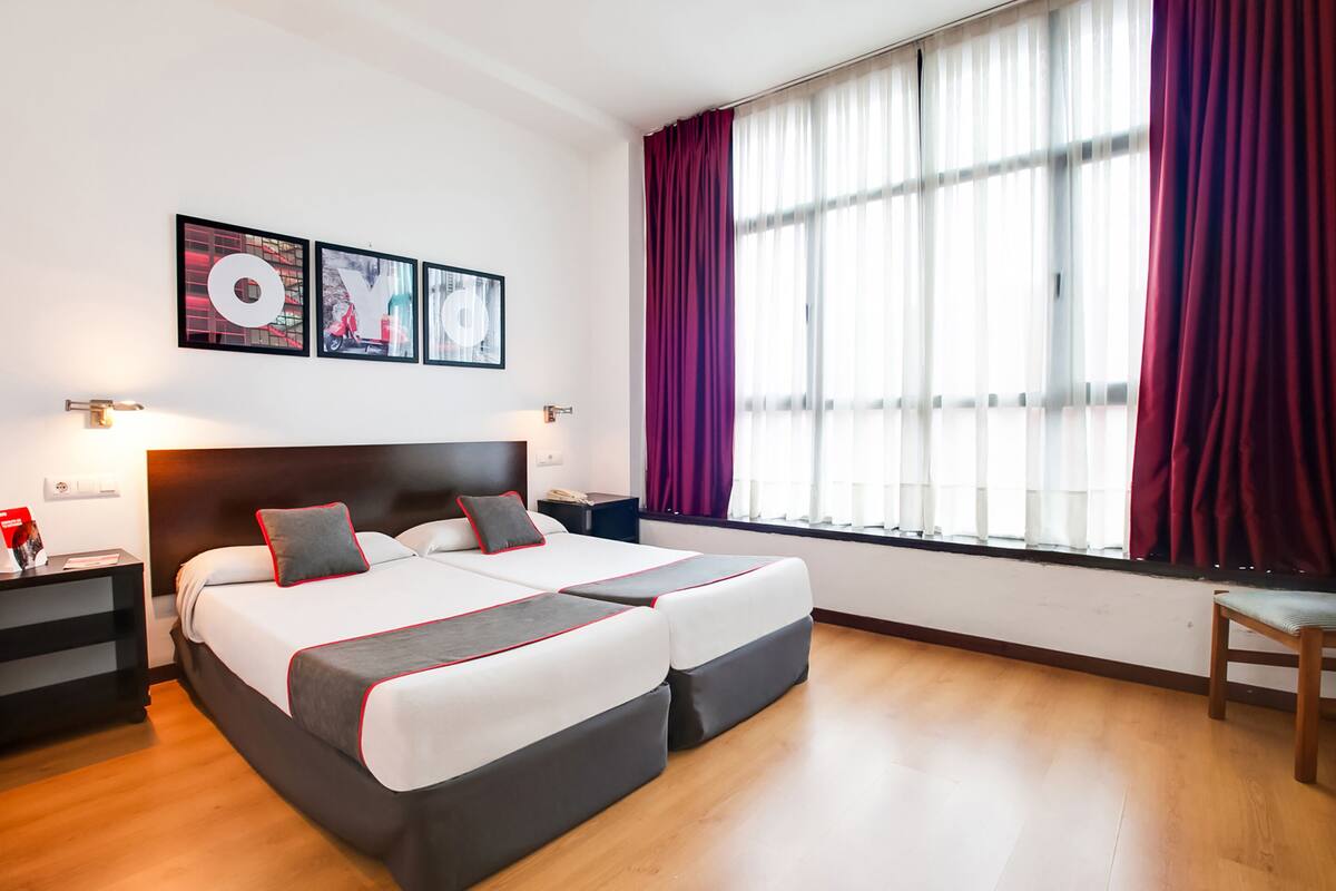 A double bed with a headboard, two bedside tables, a nightstand, a window with curtains, and wall art.