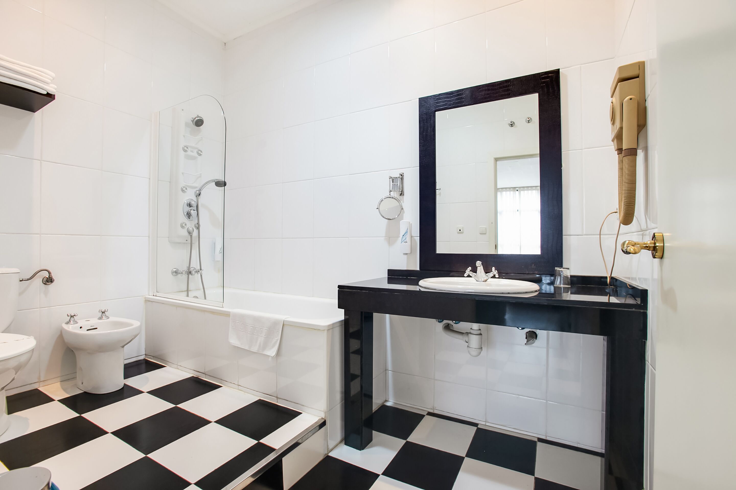 Un baño con suelo a cuadros blanco y negro, una bañera, un lavabo con espejo y un inodoro.