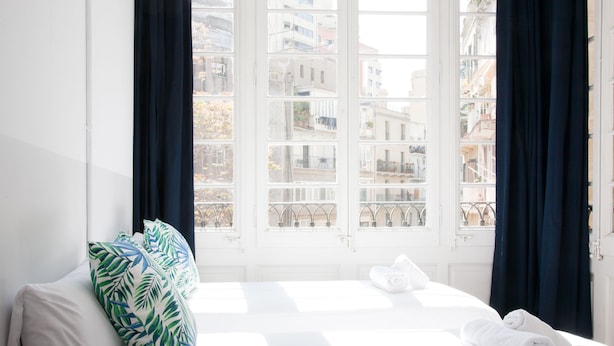 A hotel room with a bed, white curtains, and a view of buildings through a window. - My Address in Barcelona (Barcelona)