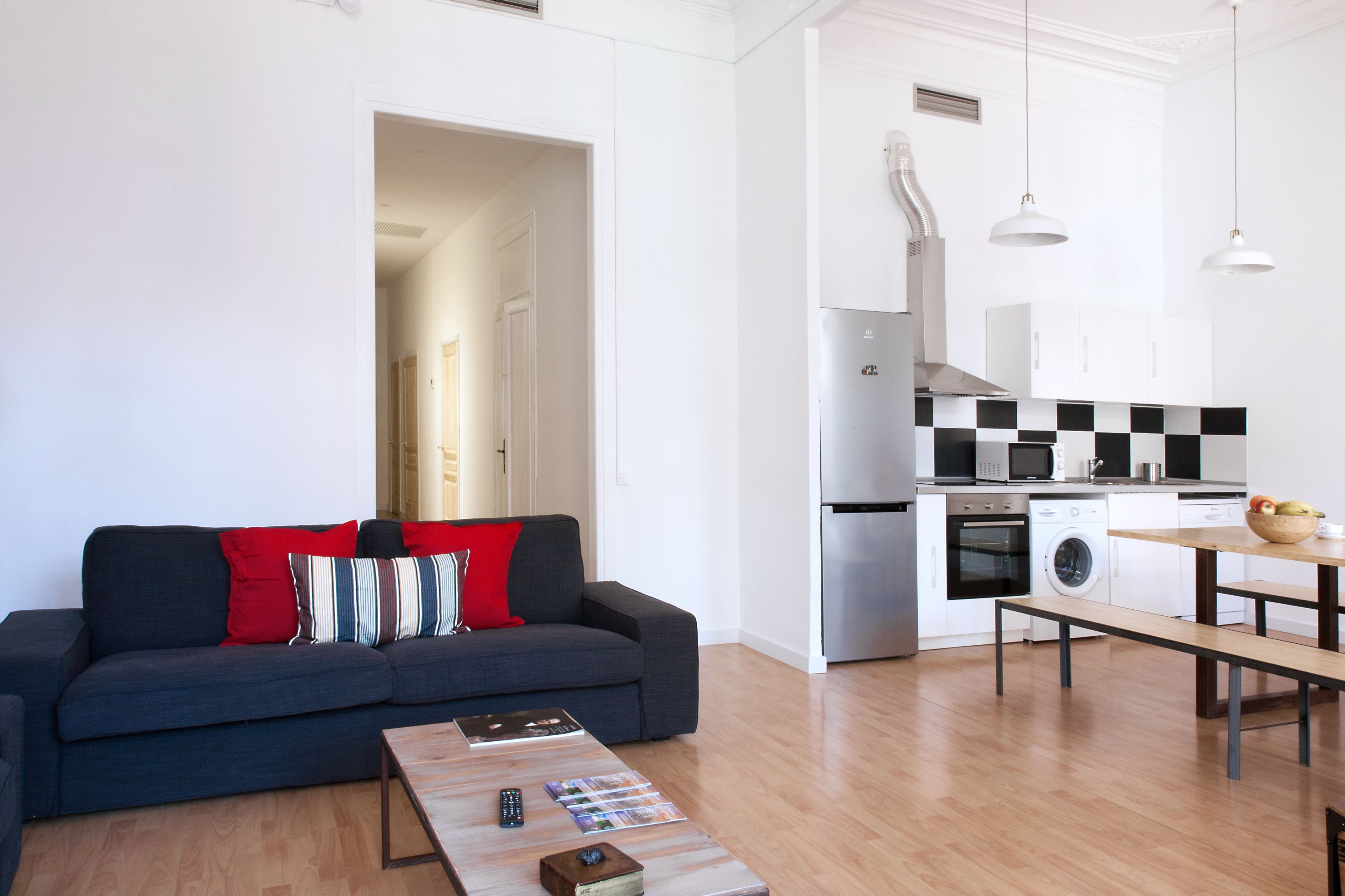A modern living room with a dark blue sofa, a wooden coffee table, and a kitchen area with white cabinets, a microwave, and a sink.