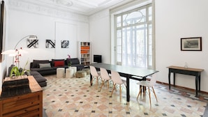 A spacious living room with a large window, a sofa, a dining table with chairs, a bookshelf, and a television. - My Address in Barcelona (Barcelona)