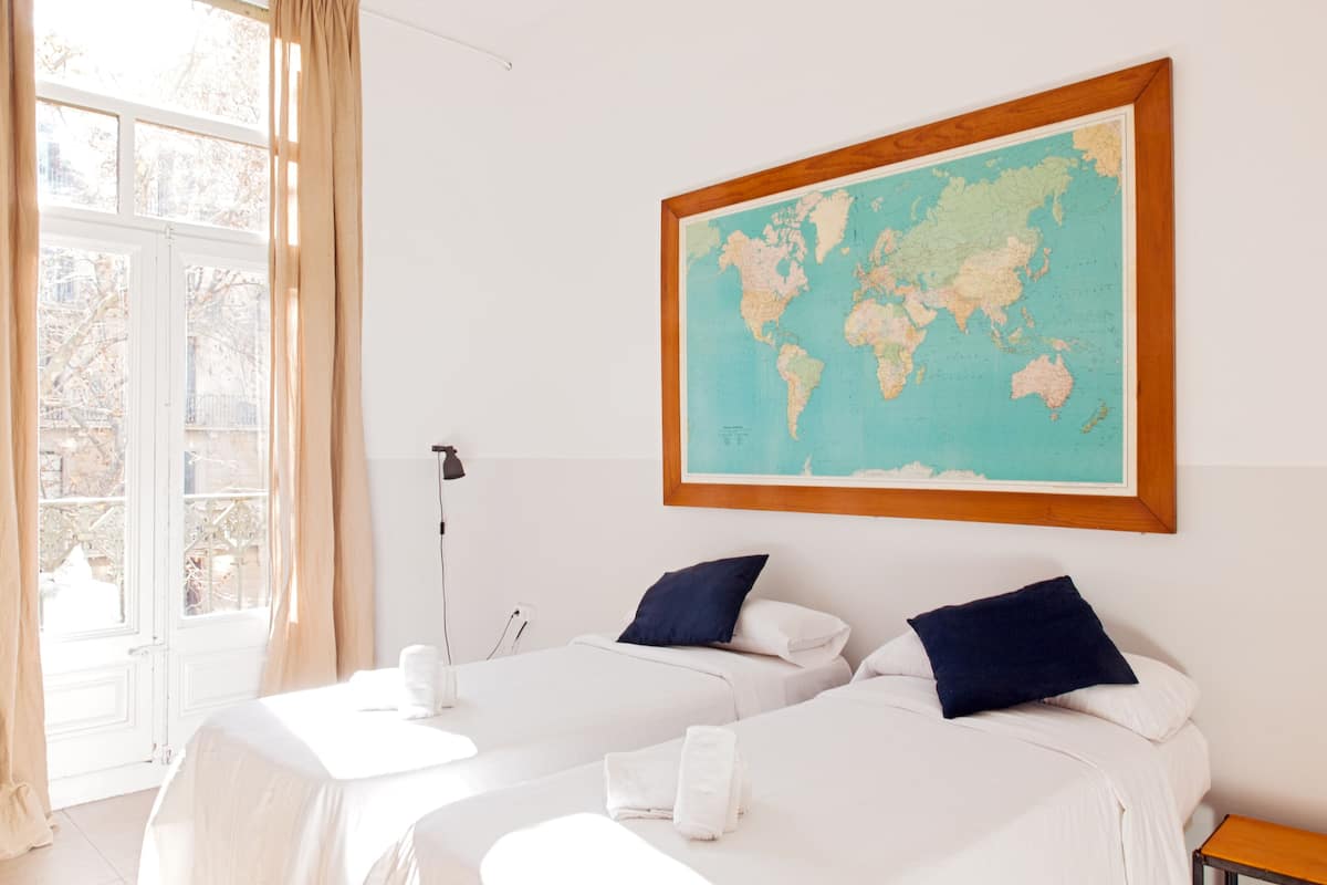A hotel room with two beds, a world map on the wall, and a window with curtains.