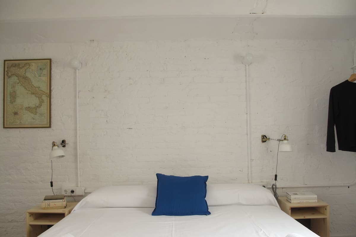 A neatly made bed with white linens and a blue pillow in a room with white brick walls. There are two bedside tables, each with a lamp and a small wooden box. A map is hanging on the wall above the bed.