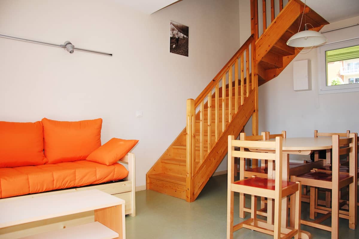A room with a wooden staircase, a wooden dining table with chairs, a white bench, and an orange sofa.