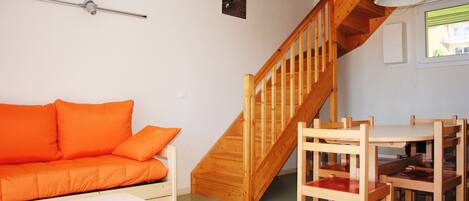 Een kamer met een houten trap, een houten eettafel met stoelen, een witte bank en een oranje banksofa.
