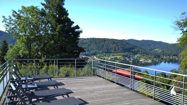 A wooden deck with lounge chairs overlooking a lake and forested hills. - Résidence Le Herbau (Gerardmer)