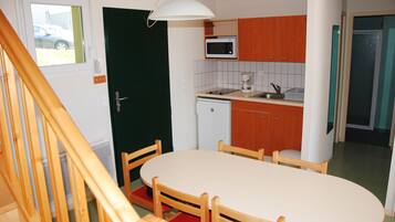 Une cuisine compacte équipée d’un four à micro-ondes, d’une machine à café et d’un petit réfrigérateur. On y trouve une table à manger avec des chaises, et un escalier en bois est visible au premier plan.