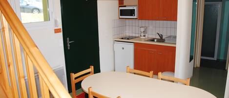 A compact kitchen with a microwave, coffee maker, and a small refrigerator. There is a dining table with chairs and a wooden staircase in the foreground.