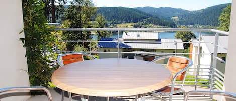 Un balcon avec une table ronde et quatre chaises, donnant sur un lac et des collines boisées.