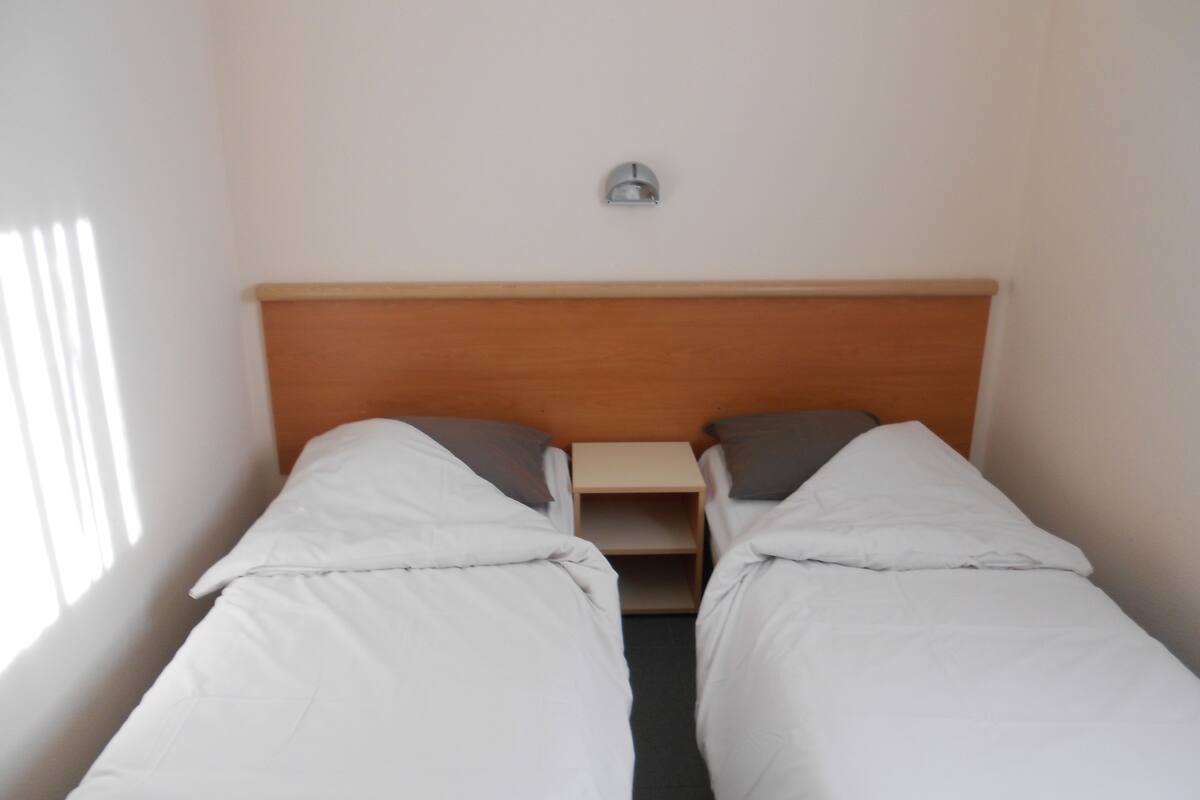A single room with two single beds, a wooden headboard, a small bedside table, and a wall-mounted light fixture.