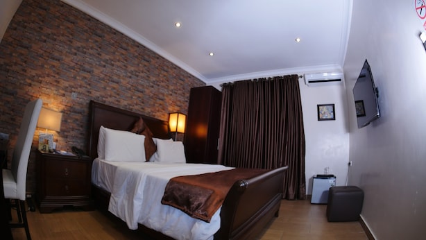 A hotel room with a brick wall, a bed with white linens, a wooden headboard, a nightstand with a lamp, a TV mounted on the wall, and a suitcase on the floor. - Fryburg Suites (Lagos)