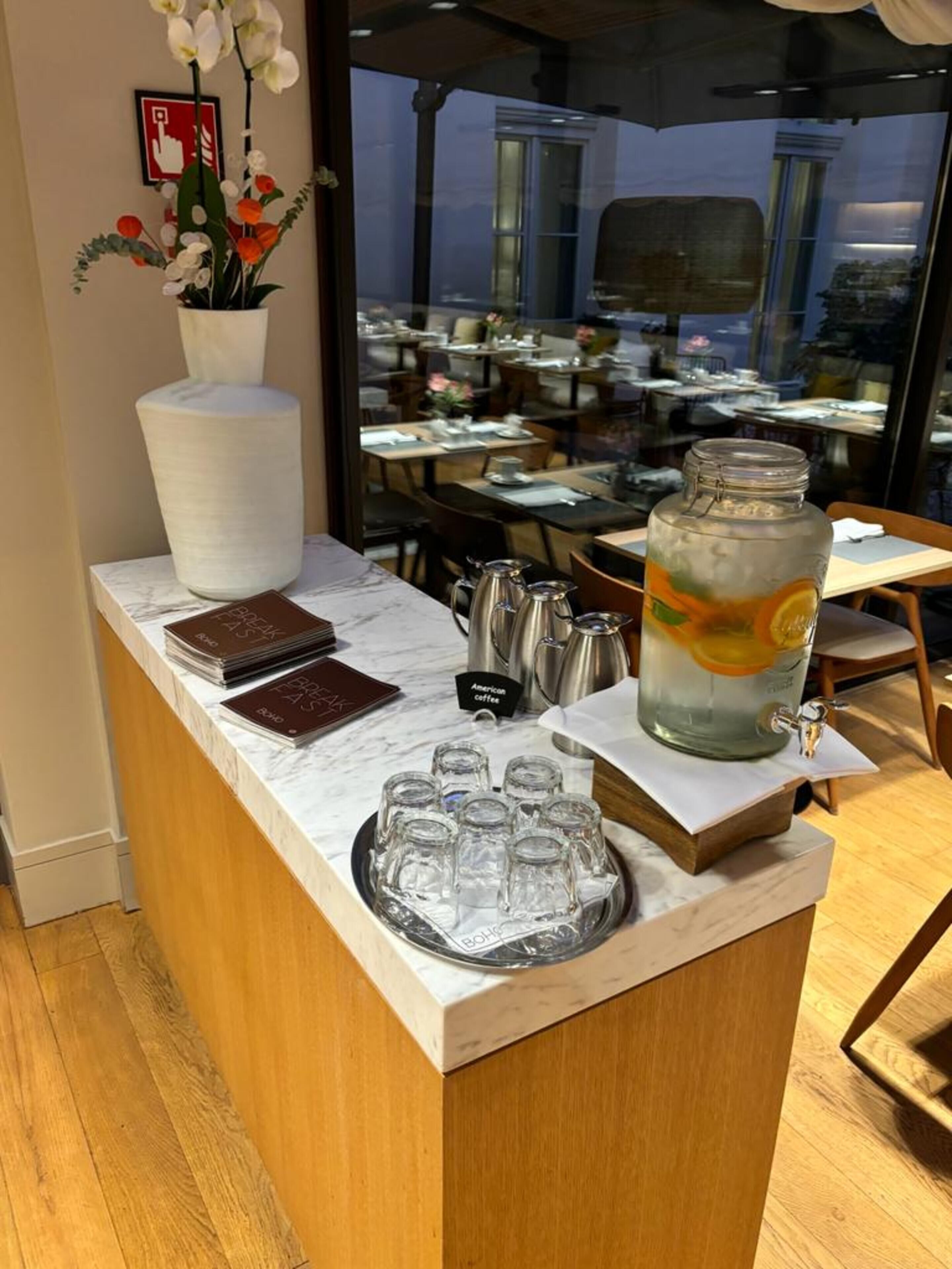 Zona de recepción de un hotel con mostrador de mármol, un florero, una estación de café y vistas al comedor a través del vidrio.