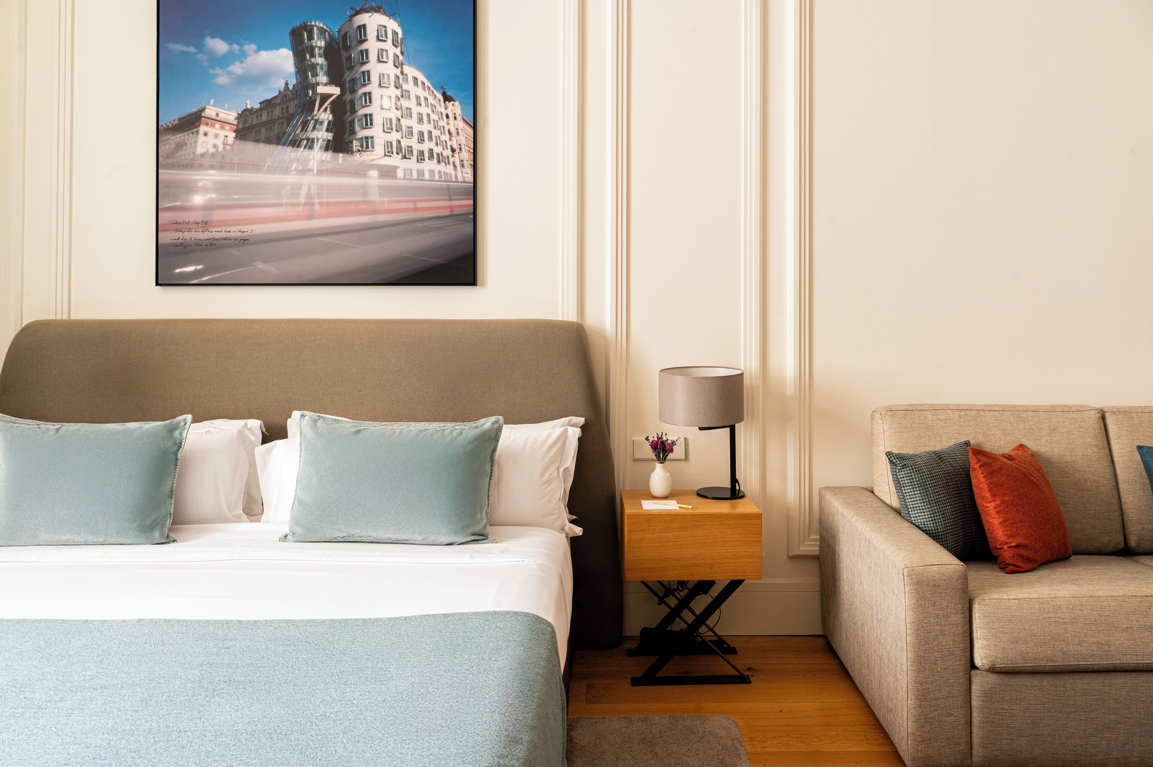 Una camera d'albergo con un letto, un comodino con una lampada, un divano e un quadro con una vista cittadina appesa al muro.