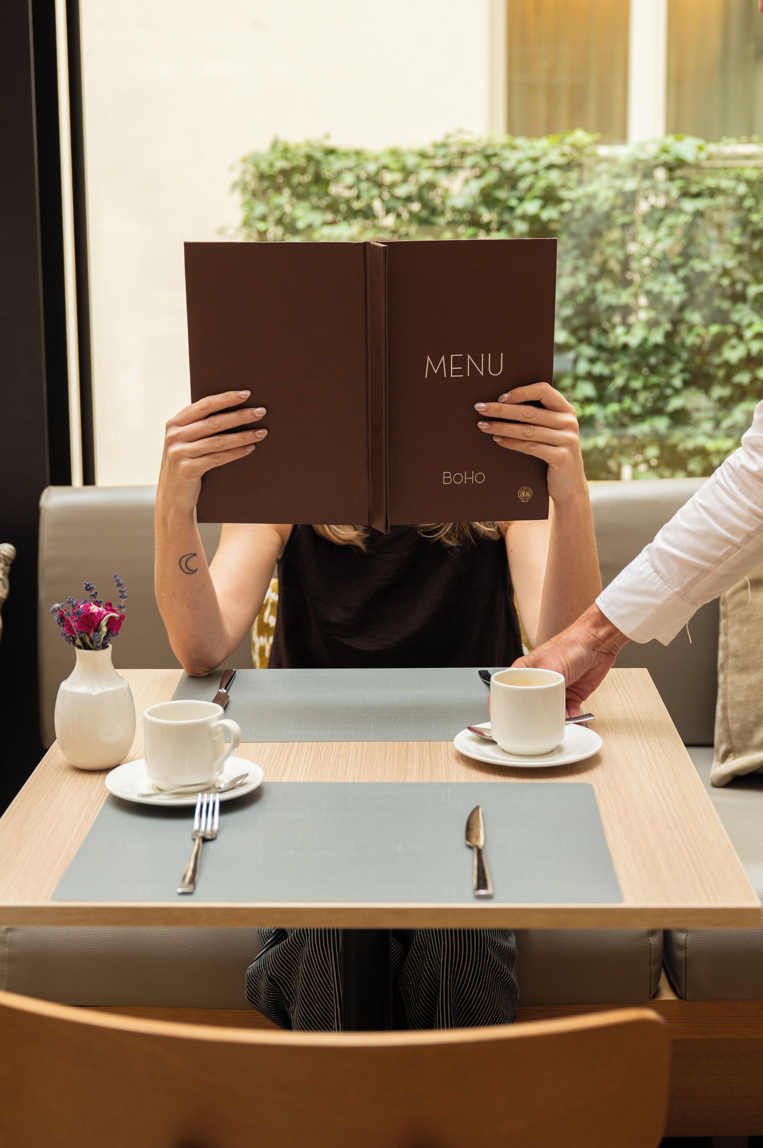 A person holding a menu at a table with a cup and saucer, a vase with flowers, and a fork and spoon on a napkin.