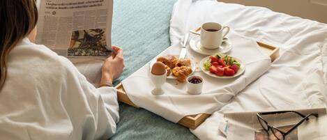 A person reading a newspaper on a bed with a tray of food and a cup of coffee.