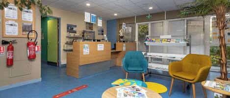 A reception area with a wooden desk, a blue and yellow chair, a fire extinguisher, and a bulletin board with notices.