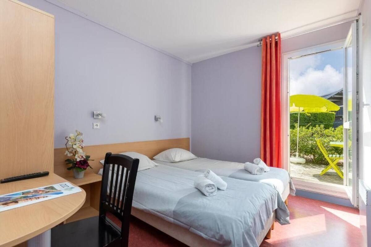 A hotel room with two beds, a desk, a chair, a small table with a vase of flowers, and a window with a view of greenery and a yellow umbrella.