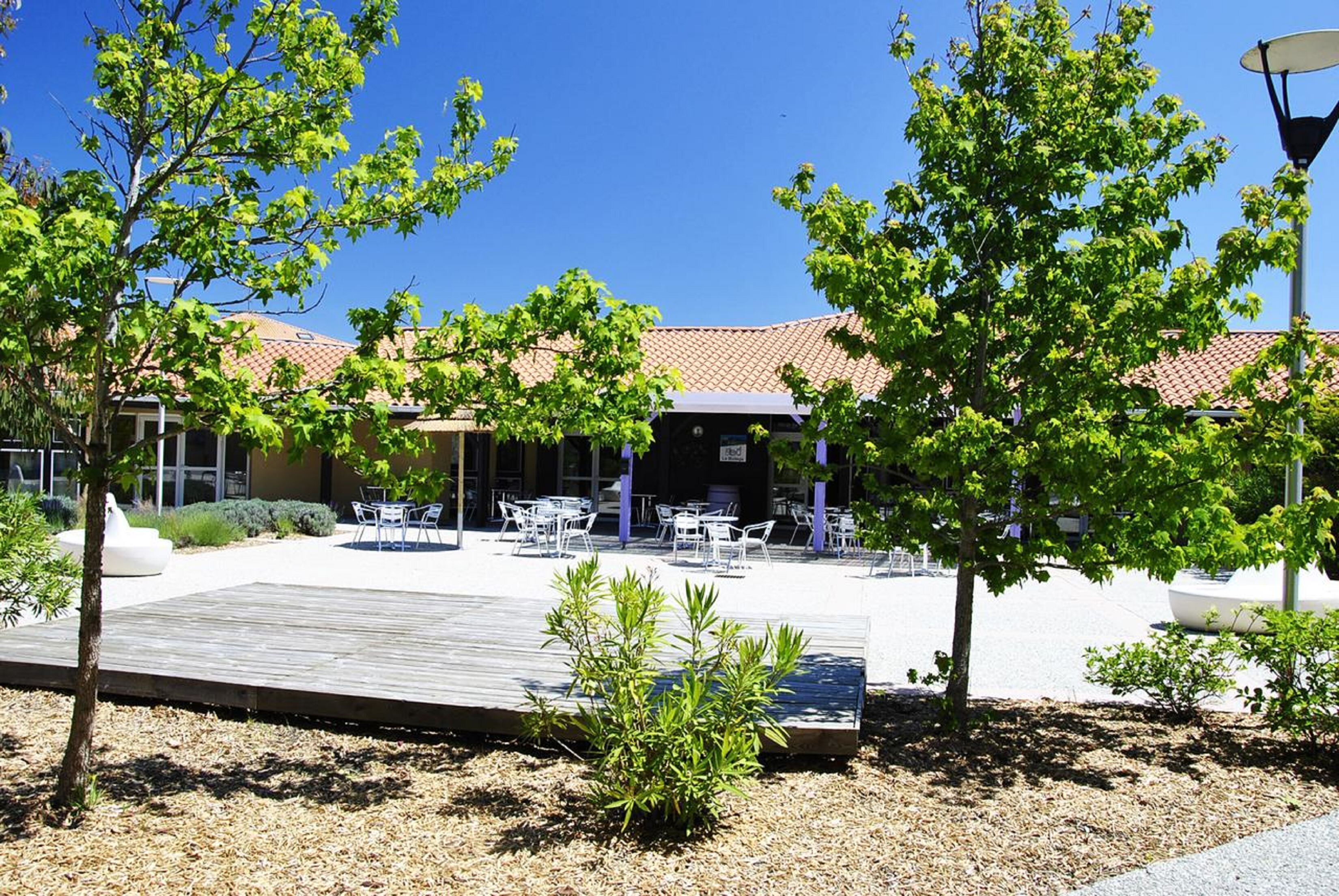 Un espace extérieur aménagé en terrasse, avec des tables et des chaises, entouré d’arbres et bordé d’un bâtiment dont le toit est recouvert de tuiles.